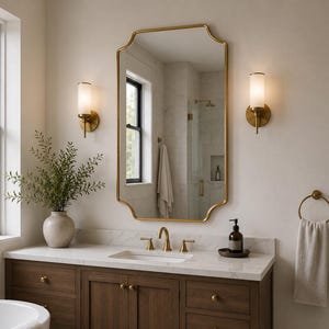 May include: A bathroom vanity featuring a dark wood cabinet with brass knobs, a white marble countertop, a gold-framed scalloped mirror, two wall-mounted cylindrical sconces, a gold faucet, and a ceramic vase with green foliage.