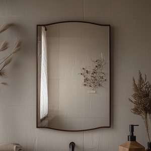 May include: A white porcelain sink with a chrome faucet, a wooden soap dispenser, and a brown glass bottle on a wooden countertop. The countertop is in front of a white tiled wall with a mirror above the sink. There are dried plants in a vase on the right side of the sink.