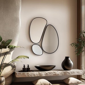 May include: A modern bathroom vanity with a black ceramic sink and a set of three black soap dispensers. The vanity is made of light grey stone and has a black, organic-shaped mirror hanging above it.