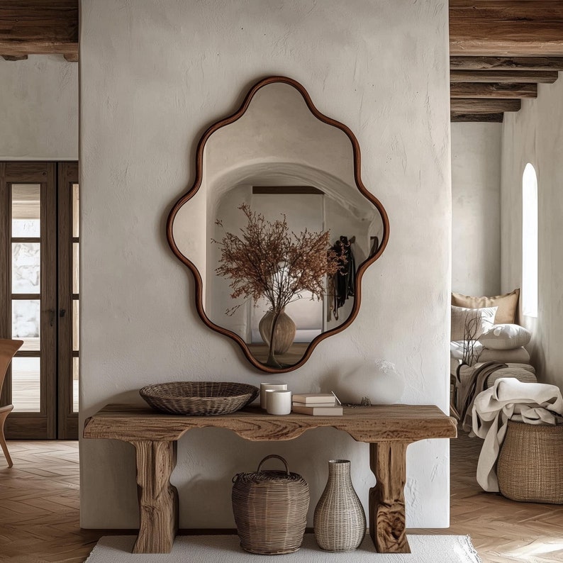 Op de afbeelding: Een houten consoletafel met een bruin blad en poten. Op de tafel staan een geweven mand, een vaas met gedroogde bloemen en twee kaarsen. De tafel staat voor een witte muur met een grote, sierlijke spiegel.