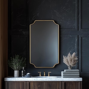 May include: A gold framed mirror with a beveled edge hangs above a white marble countertop with a double sink and gold faucets. The countertop is in front of a dark gray wall with wood paneling. There are two plants and a bottle of soap on the countertop.