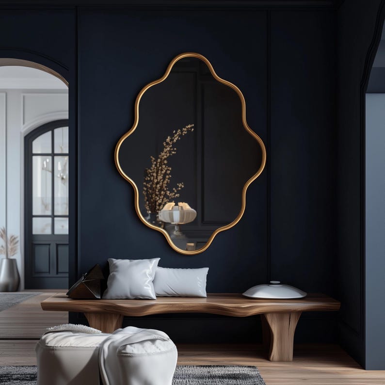 Op de afbeelding: Een houten bank met een wit kussen en een wit deken staat voor een donkerblauwe muur. Een gouden, sierlijke spiegel met een golvende lijst hangt aan de muur boven de bank. De spiegel reflecteert een boeket gedroogde bloemen.