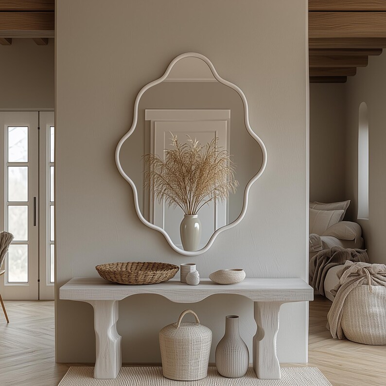 Op de afbeelding: Een witte spiegel met golvende rand hangt boven een witte consoletafel met een geweven mand, twee witte keramische vazen en een witte keramische kom. De tafel staat in een kamer met een lichte houten vloer en een deur die naar een andere kamer leidt.
