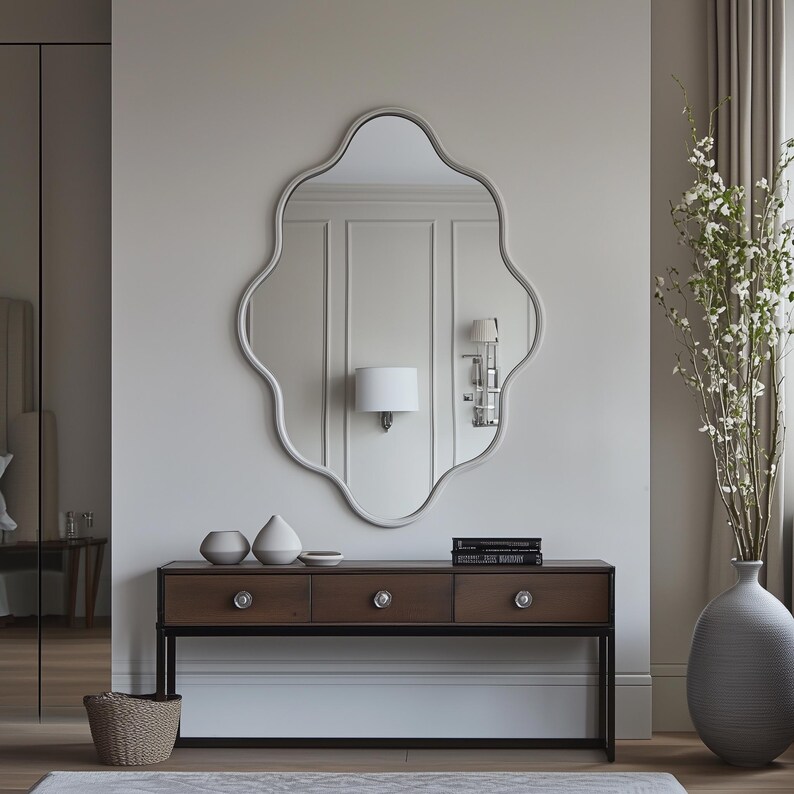 Op de afbeelding: Een witte, sierlijke spiegel met een gegolfd frame hangt boven een donkerbruine consoletafel met drie laden. De tafel heeft zwarte metalen poten en zilveren ladegrepen. Twee witte keramische vazen en een klein dienblad staan op de tafel. Een hoge, grijze keramische vaas met een witte bloemenarrangement staat rechts in de afbeelding.