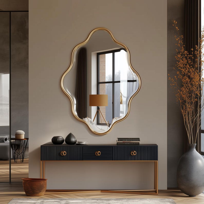 Op de afbeelding: Een golvende spiegel met een gouden frame hangt boven een zwarte consoletafel met drie laden. De tafel heeft gouden accenten en staat op een houten vloer.