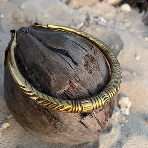 May include: A gold-coloured, patterned bracelet sits on a weathered coconut. The bracelet has etched designs and a curved, open design. The background shows sand and driftwood, suggesting a beach setting.