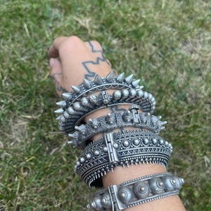 Może przedstawiać: Kolekcja srebrnych bransoletek z metalu o różnych wzorach, w tym kolczastych, ćwiekowanych i wzorzystych teksturach. Bransoletki są ułożone na nadgarstku na tle zielonej trawy.