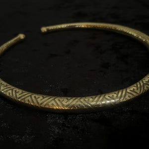 May include: A gold-colored, open-ended collar necklace with an intricate geometric pattern etched along its surface. The necklace is circular and rests on a dark background, highlighting its metallic sheen.