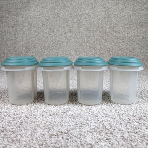 May include: Four small, cylindrical, translucent plastic containers arranged in a row. Each container features a removable, circular, teal-colored lid with a slightly raised, ridged edge. The containers have a clear, semi-transparent body.