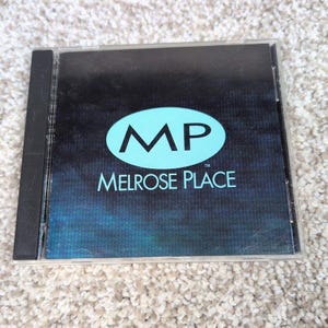 May include: A CD case with a black cover featuring a teal oval with the letters "MP" inside and the words "Melrose Place" below. The background is a dark blue gradient.