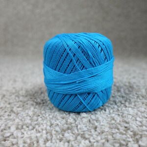 May include: A spherical ball of bright cyan blue crochet thread wound in a crisscross pattern. The thread is tightly coiled, creating a textured surface with visible individual strands forming a dense, rounded shape for crafting projects.