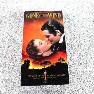 May include: A VHS tape of the movie "Gone With The Wind." The cover features a romantic image of two people kissing, set against an orange and black background. The text "Gone With The Wind" is at the top, and the words "Winner Of 10 Academy Awards" are at the bottom.