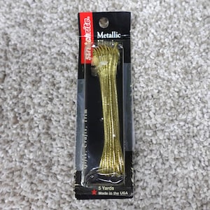 May include: A package of Stretchrite brand metallic gold cord trim. The packaging is black with white text and includes the label 5 Yards Made in the USA. The gold cord is coiled and visible through a clear plastic window on the front of the card.