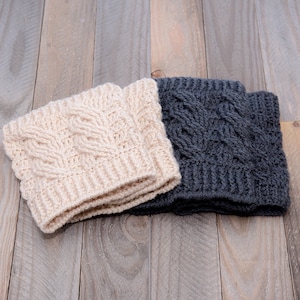 Könnte beinhalten: Zwei Paar gehäkelte Beinstulpen, eine in Beige und eine in Grau, mit einem Zopfmuster. Die Beinstulpen sind gefaltet und auf einer Holzoberfläche platziert.