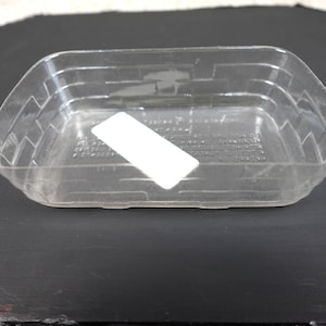May include: A clear, rectangular plastic container with a basket-weave texture molded into the sides. The container features a smooth, white rectangular label adhered to the interior base, partially obscuring faint, embossed text on the bottom.