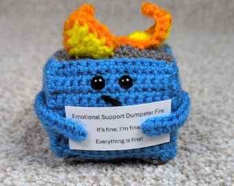 May include: A blue crochet cube with black button eyes and a small black mouth. The cube has orange and yellow flames on top and is holding a sign that reads "Emotional Support Dumpster Fire. It's fine. I'm fine, Everything is fine!"