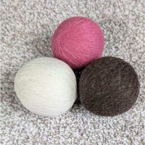 May include: Three wool balls in a close-up shot. One is a dusty rose color, another is off-white, and the third is a dark brown. The balls are round and appear to be made of soft, textured wool. The background is a light, textured surface.