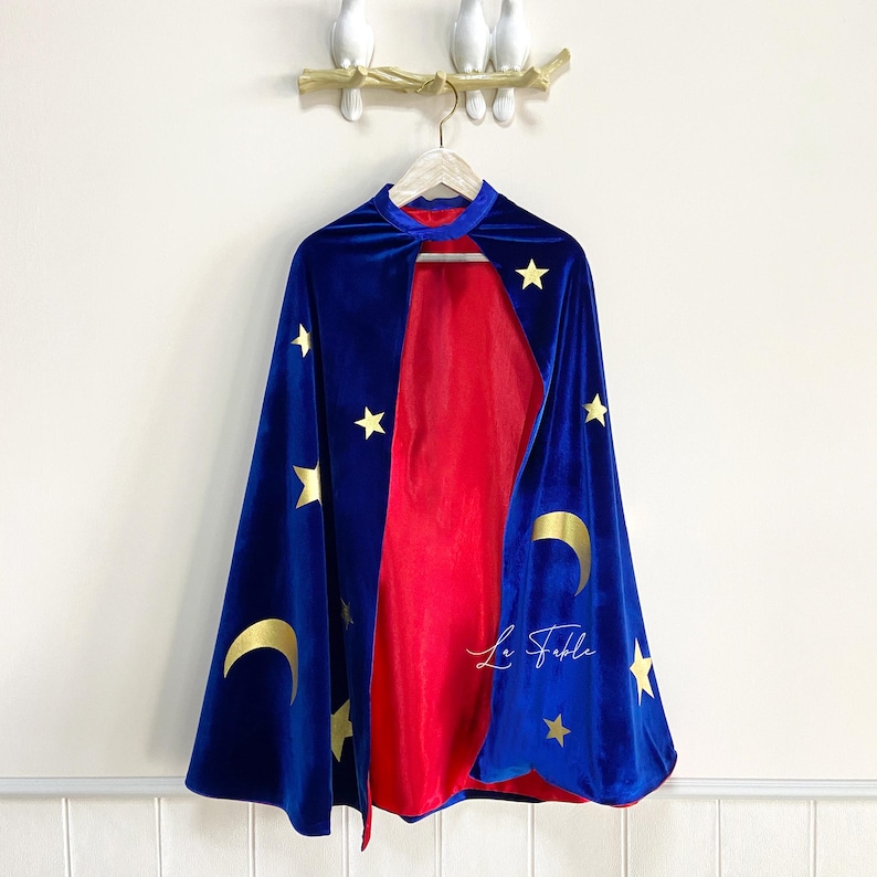 May include: A blue velvet cape with gold stars and crescent moons. The cape has a red lining and is hanging on a hanger.