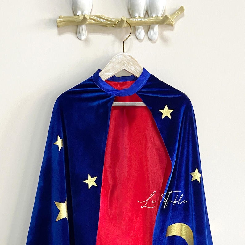 May include: A blue velvet cape with gold stars and a crescent moon design. The cape has a red lining and is hanging on a wooden hanger.