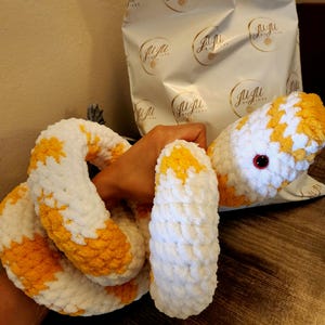 Puede incluir: Un juguete de serpiente de crochet en blanco y amarillo. La serpiente está enroscada y tiene un ojo rojo.