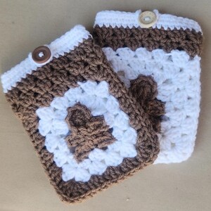May include: Two handmade crochet cup cozies. Each cozy features a brown and white granny square design with a brown flower in the center. The top edge is white with a brown button closure. These cozies are perfect for keeping your hands safe from hot beverages.