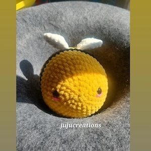 May include: A crocheted bee plush toy with yellow and black stripes, white wings, and a cute face. The bee is sitting on a grey surface. _jujucreations_