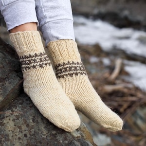 Puede incluir: Un par de calcetines de punto de color crema con patrones geom&eacute;tricos marrones. Los calcetines est&aacute;n puestos en los pies de una persona y descansan sobre una roca.