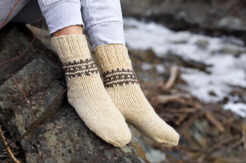 Puede incluir: Un par de calcetines de punto de color crema con patrones geom&eacute;tricos marrones. Los calcetines est&aacute;n puestos en los pies de una persona y descansan sobre una roca.
