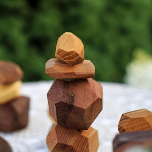 May include: Wooden geometric stacking toy in various shades of brown. The pieces are faceted and stacked to create a tower. The background is a blurred green and white.