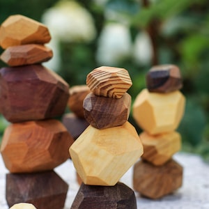 Puede incluir: Piedras apilables de madera en varios tonos de marrón y beige. Las formas geométricas están apiladas para crear torres. La veta natural de la madera es visible. El fondo está borroso con vegetación.