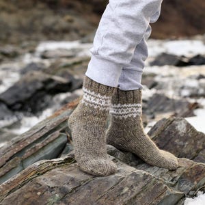 Puede incluir: Un par de calcetines de lana marr&oacute;n con detalles blancos. Los calcetines est&aacute;n puestos en los pies de una persona y descansan sobre una superficie rocosa.