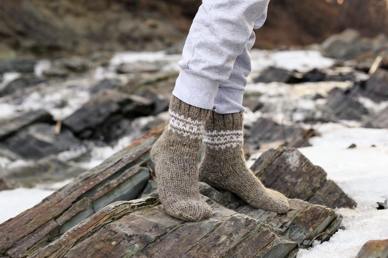 Puede incluir: Un par de calcetines de lana marr&oacute;n con detalles blancos. Los calcetines est&aacute;n puestos en los pies de una persona y descansan sobre una superficie rocosa.