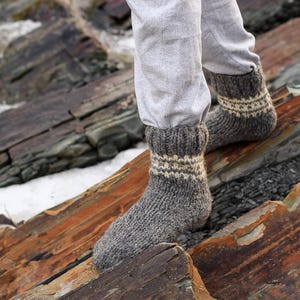 Puede incluir: Un par de calcetines de lana grises con rayas blancas. Los calcetines est&aacute;n puestos en los pies de una persona y est&aacute;n de pie sobre una superficie rocosa.