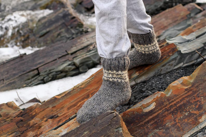Puede incluir: Un par de calcetines de lana grises con rayas blancas. Los calcetines est&aacute;n puestos en los pies de una persona y est&aacute;n de pie sobre una superficie rocosa.