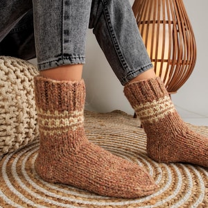 May include: A pair of brown and cream knitted socks with a geometric pattern. The socks are worn by a person sitting on a woven rug.