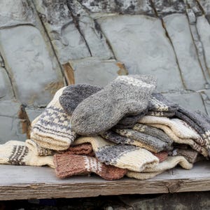May include: A pile of hand-knitted wool socks in various colors, including cream, gray, and brown. The socks feature patterned designs and are arranged on a weathered wooden surface against a backdrop of gray rock.