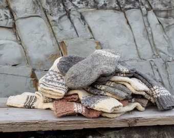 May include: A pile of hand-knitted wool socks in various colors, including cream, gray, and brown. The socks feature patterned designs and are arranged on a weathered wooden surface against a backdrop of gray rock.