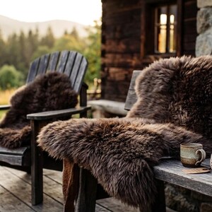 May include: Two wooden Adirondack chairs and a wooden bench are adorned with brown sheepskin throws. A rolled blanket and a mug sit on the bench. The scene is set outdoors with a backdrop of trees and a rustic cabin.