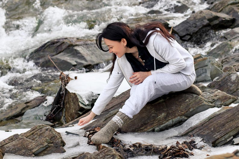 Puede incluir: Una mujer con una sudadera con capucha blanca y pantalones blancos se sienta en una roca junto a un arroyo. Est&aacute; mirando algo en su mano. El arroyo est&aacute; parcialmente congelado y hay nieve en las rocas.