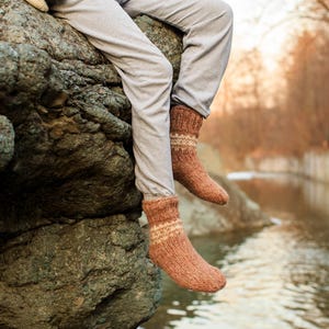 May include: A person wearing brown and white knitted socks sits on a large rock with a river in the background.