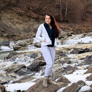Puede incluir: Una mujer con una chaqueta blanca y pantalones grises se encuentra de pie en un arroyo rocoso. Lleva calcetines de lana marrones y tiene el pelo largo y casta&ntilde;o.