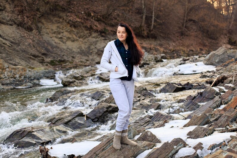 Puede incluir: Una mujer con una chaqueta blanca y pantalones grises se encuentra de pie en un arroyo rocoso. Lleva calcetines de lana marrones y tiene el pelo largo y casta&ntilde;o.