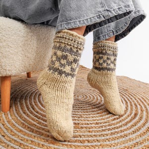 May include: A pair of beige wool socks with a grey geometric pattern. The socks are worn by a person sitting on a white fluffy stool. The stool is on a brown and white circular rug.