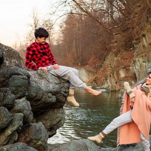 Puede incluir: Un joven con una camisa de cuadros rojos y negros se sienta en una gran roca junto a un r&iacute;o. Sonr&iacute;e y mira a una joven que est&aacute; sentada en una roca en el r&iacute;o. La mujer lleva una manta rosa y tiene los brazos alrededor de s&iacute; misma. El r&iacute;o fluye detr&aacute;s de ellos y hay &aacute;rboles en el fondo.