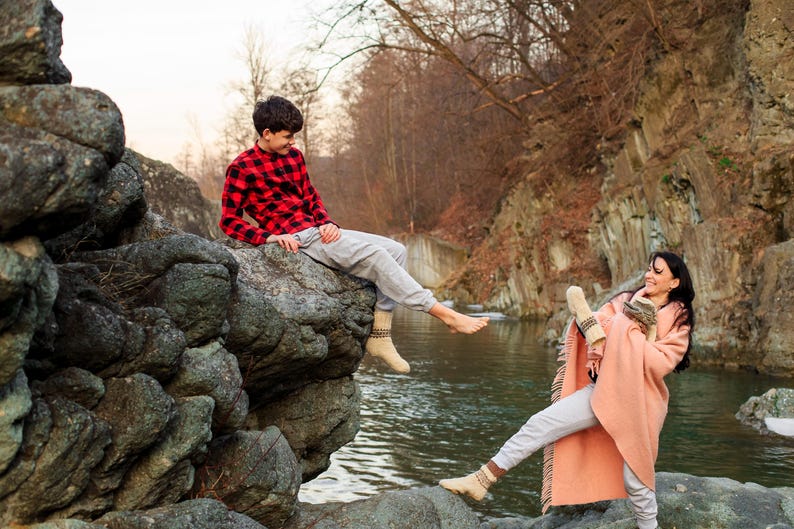 Puede incluir: Un joven con una camisa de cuadros rojos y negros se sienta en una gran roca junto a un r&iacute;o. Sonr&iacute;e y mira a una joven que est&aacute; sentada en una roca en el r&iacute;o. La mujer lleva una manta rosa y tiene los brazos alrededor de s&iacute; misma. El r&iacute;o fluye detr&aacute;s de ellos y hay &aacute;rboles en el fondo.