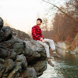 Puede incluir: Una persona joven con una camisa de cuadros rojos y negros y pantalones beige se sienta en una gran roca junto a un arroyo. La persona est&aacute; mirando hacia arriba y sonriendo. El fondo es un bosque con &aacute;rboles y rocas.