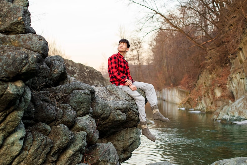 Puede incluir: Una persona joven con una camisa de cuadros rojos y negros y pantalones beige se sienta en una gran roca junto a un arroyo. La persona est&aacute; mirando hacia arriba y sonriendo. El fondo es un bosque con &aacute;rboles y rocas.