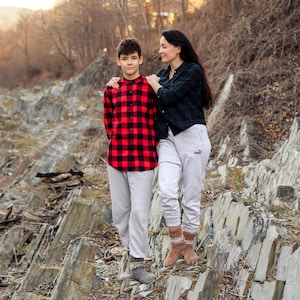 Puede incluir: Una persona joven con una camisa a cuadros rojos y negros y pantalones blancos se para junto a un adulto con una camisa negra y pantalones blancos. Est&aacute;n parados en una superficie rocosa.
