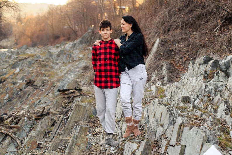 Puede incluir: Una persona joven con una camisa a cuadros rojos y negros y pantalones blancos se para junto a un adulto con una camisa negra y pantalones blancos. Est&aacute;n parados en una superficie rocosa.