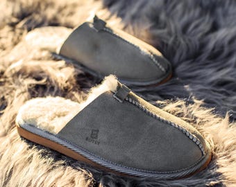 May include: A pair of gray suede slippers with a soft, white interior lining. The slippers have a stitched design around the edges and a small strap detail. The slippers are on a gray, fluffy surface.